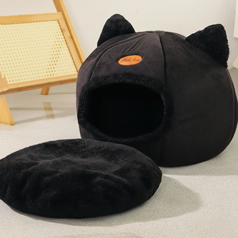 Black cat-shaped pet bed on a light gray floor with a wooden chair in the background.